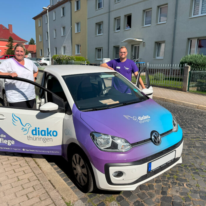 Zwei lächelnde Teammitglieder des Diako Pflegedienstes Eisenach stehen am Dienstauto mit Diako Thüringen Beklebung
