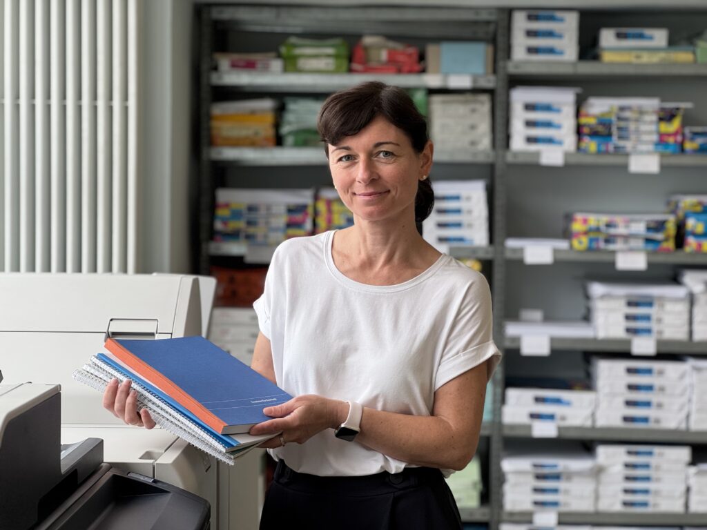 Mitarbeiterin unserer Diakopie steht neben den Druckmaschinen und zeigt Druckbeispiele